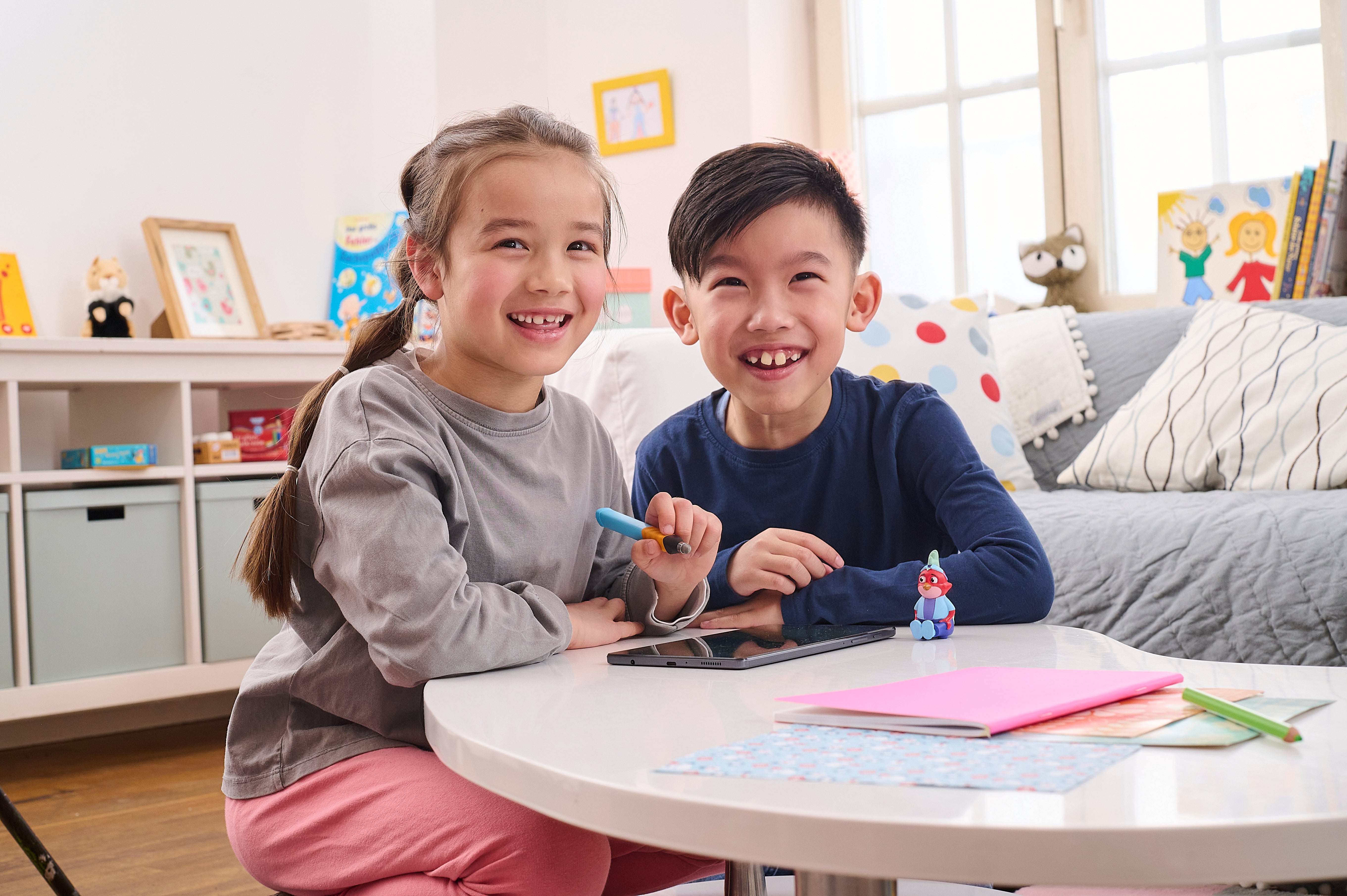 Zwei lächelnde Kinder sitzen in einem hellen, gemütlichen Wohnzimmer an einem Tisch und malen mit Filzstiften.