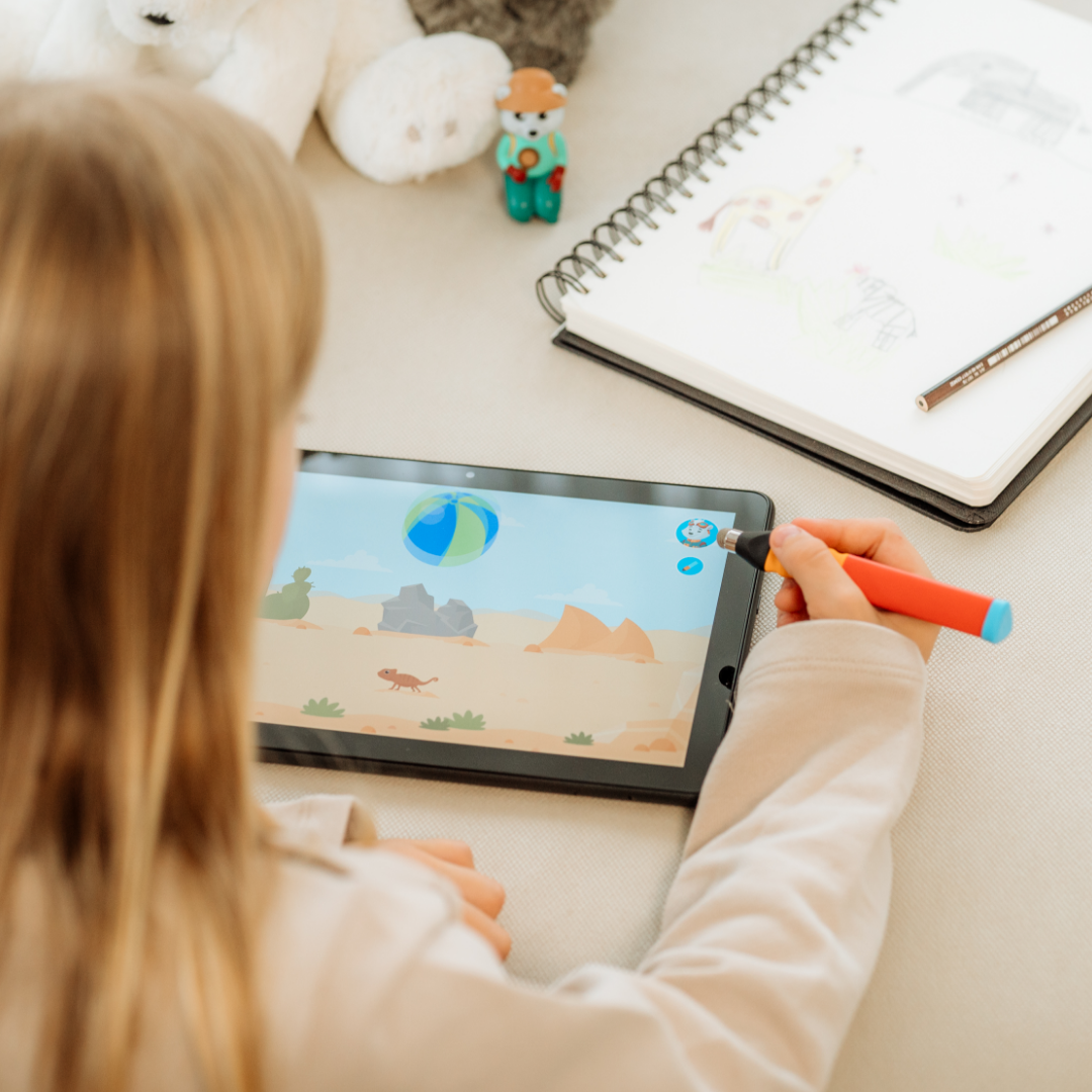 Ein Kind benutzt einen Stift auf einem Tablet mit einer Strandszene und erkundet Die 3 neuesten Lernwelten von Edurino DE für spielerisches Uhr-Erlernen, während in der Nähe ein Skizzenbuch und ein Spielzeug Konzentration trainieren helfen.