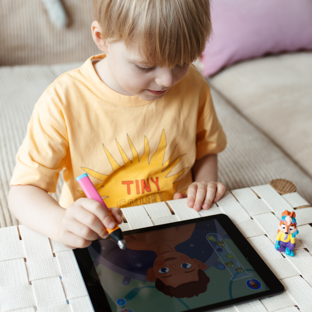 Ein Kind erforscht mit Ari "Meine Körperteile" von Edurino DE seine Sinne mit einem Stift auf einem Tablet, trägt ein gelbes Tiny-Shirt und hat eine Spielzeugfigur neben sich auf dem Tisch.