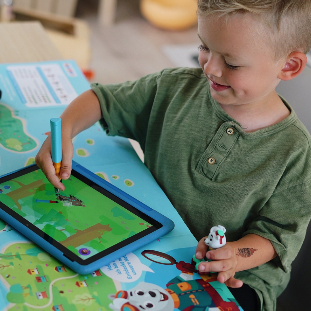 Ein lächelnder Junge benutzt einen Stift auf einem Tablet mit Asa "Unsere Natur" von Edurino DE und hält einen kleinen Spielzeughund, während eine bunte Karte mit heimischen Tieren und Pflanzen auf dem Tisch ausgebreitet ist.