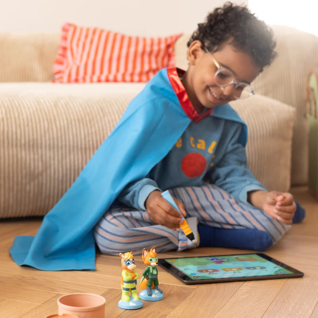 Ein lächelndes Kind in einem blauen Umhang spielt mit Mika "Wörter & Sätze" von Edurino DE auf einem iPad, umgeben von Spielzeugfiguren auf dem Wohnzimmerboden.