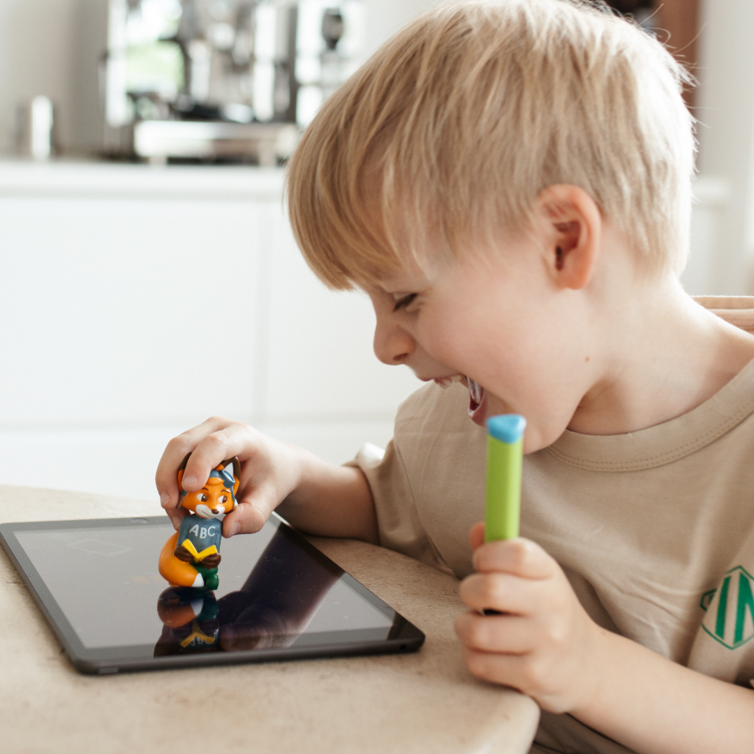 Ein lächelnder Junge benutzt das Edurino DE Mika "Buchstaben Lesen & Schreiben" - Stufe 2 auf seinem Tablet, spielt mit einer Fuchsfigur und hält einen grünen Marker in der Hand, während er sich mit Buchstaben und Schreiben beschäftigt.