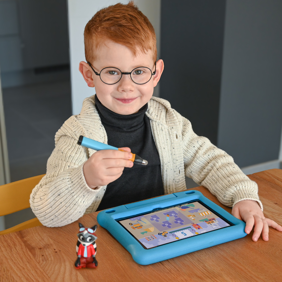 Ein lächelnder Junge mit Brille benutzt an einem Tisch einen Stift auf einem Tablett, neben ihm das Edurino DE Robin "Zahlen & Mengen" - Stufe 1 Waschbär-Spielzeug.