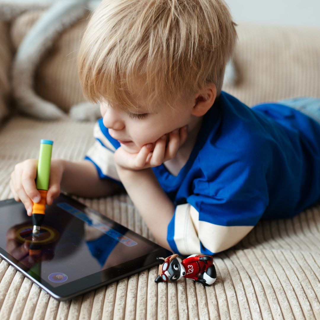 Ein kleines Kind in einem blauen Hemd benutzt Robin "Erstes Rechnen bis 10" - Stufe 2 von Edurino DE mit einem Stift auf einem Tablet, das auf einer beigen Couch neben einer Spielzeugfigur liegt, während es sich spielerisch mit Zahlen und einfachen Matheaufgaben beschäftigt.