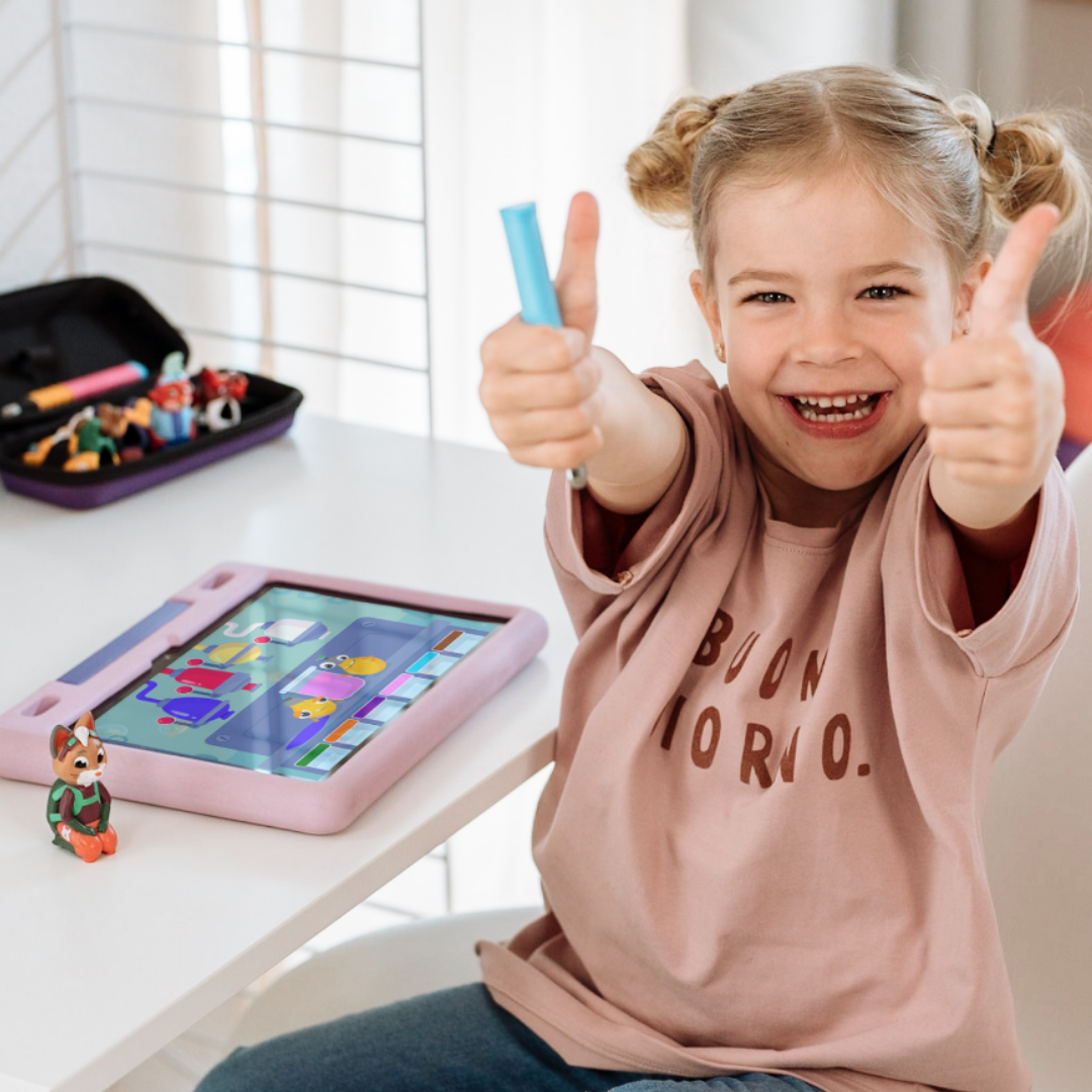 Ein lächelndes Mädchen an ihrem Schreibtisch zeigt zwei Daumen nach oben, während sie das Yuki "Kreativität & Malen" Set von Edurino DE, ein Tablet und Spielzeug vor sich hat.