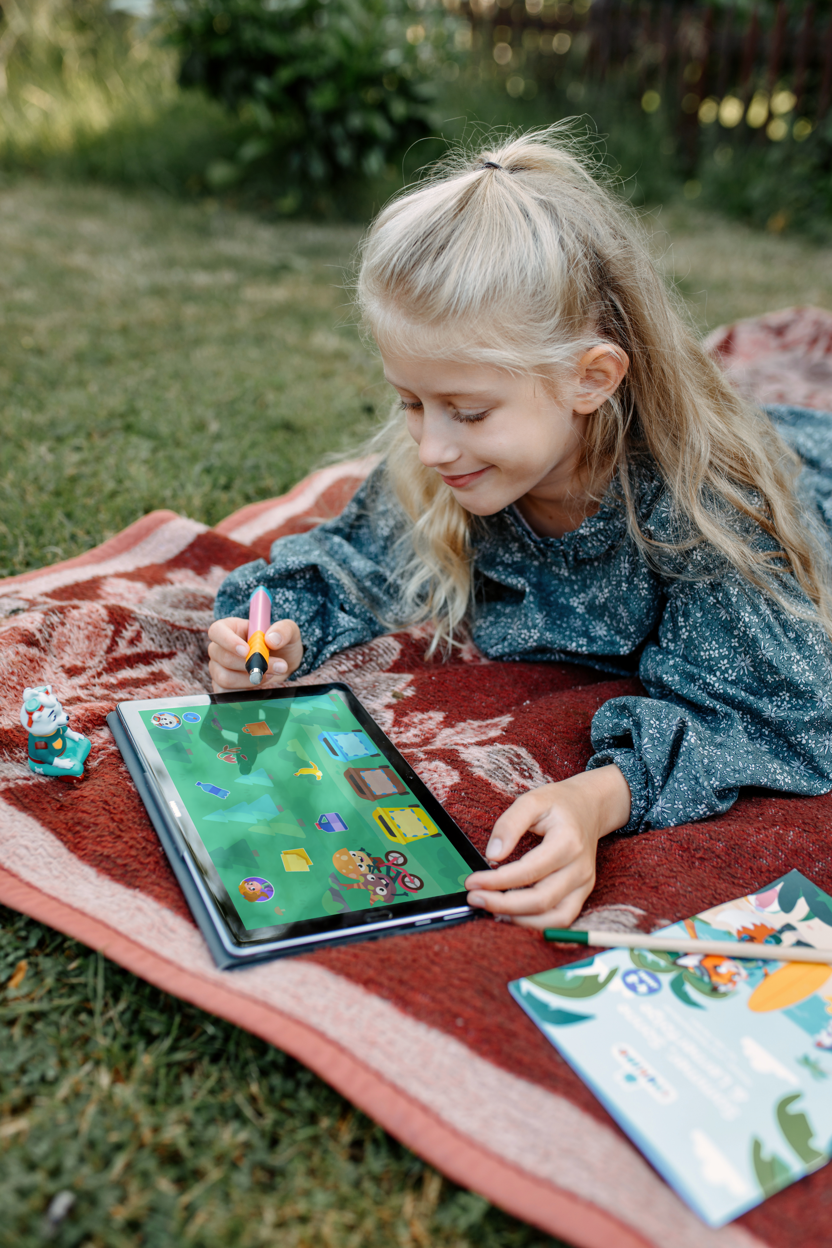 Mdchen liegt mit einem Tablet auf einer Decke im Garten, auf dem Tablet das Spiel zur Mülltrennung